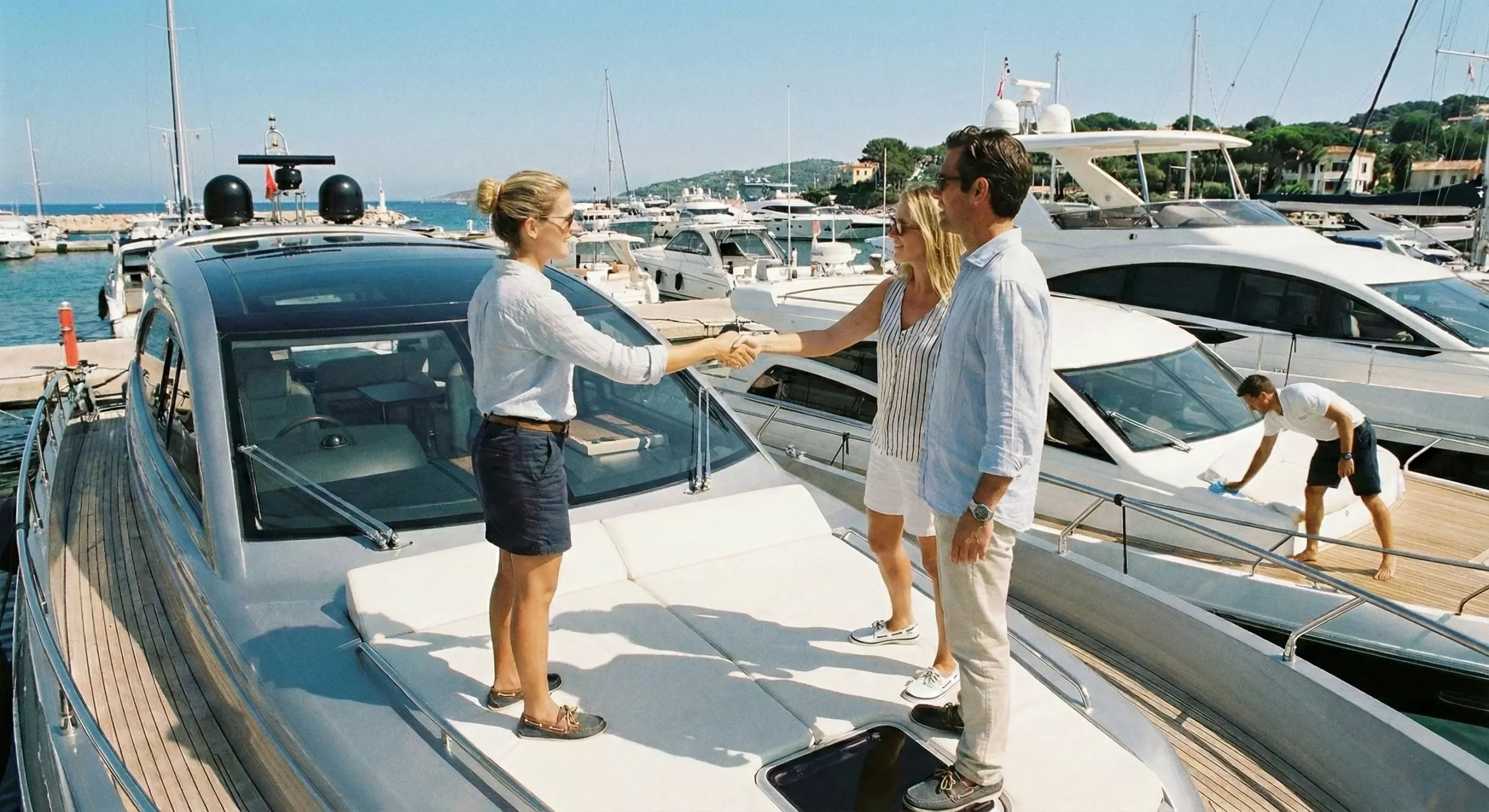 Güneşli bir marinada, lüks bir motor yatın güvertesinde, üniformalı kadın personel ile bir çiftin, denize açılmadan önceki zorunlu güvenlik brifingi ve teslimat prosedürlerinin ardından el sıkıştığı anın fotoğrafı.